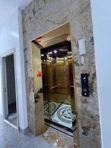 Thang máy Việt Yên tại nhà anh Phước, My Điền — cửa tầng inox titan vàng gương, tường sảnh ốp đá granite vân nâu
