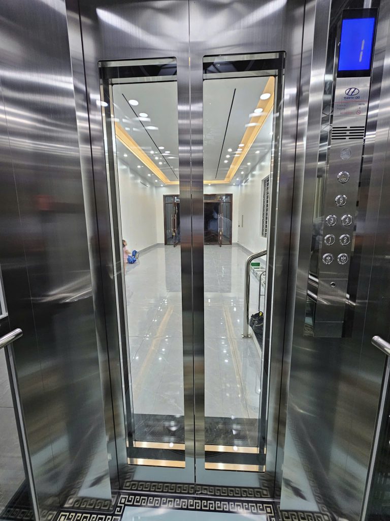 Cửa cabin thang máy kính có ô kính lớn nhìn xuyên ra sảnh, cabin inox xước và tay vịn inox
