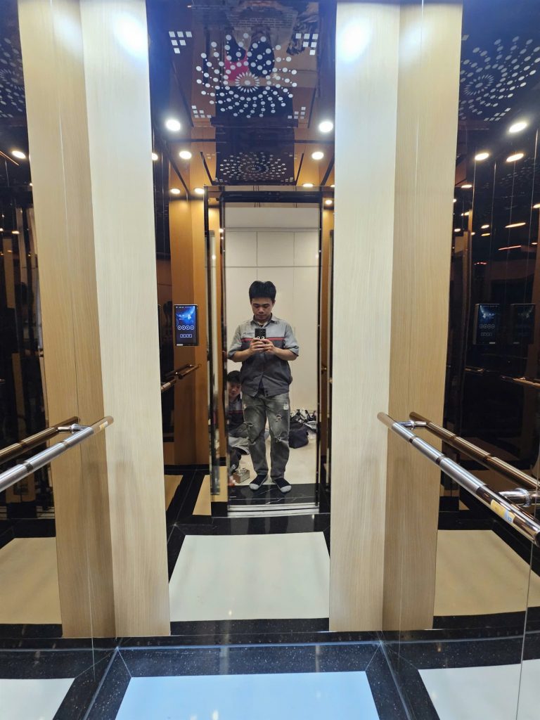Cửa tầng thang máy kính inox vàng hồng mở, nhìn vào cabin ốp vân gỗ sáng